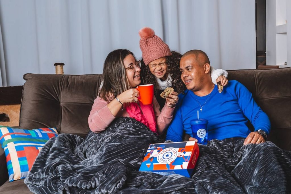Mulher toma chocolate quente sentada no sofá ao lado de homem, ambos cobertos por uma manta. Eles são abraçados por uma criança de blusa de frio e touca