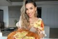 Mulher sorri enquanto segura tábua de pizza em uma mão e pedaço de pizza em outra