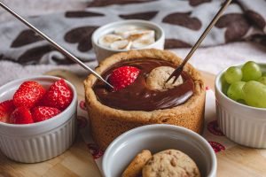 Vasilha feita de cookie com ganache de chocolate dentro e potes de porcelana ao redor, com frutas, cookies e marshmallows