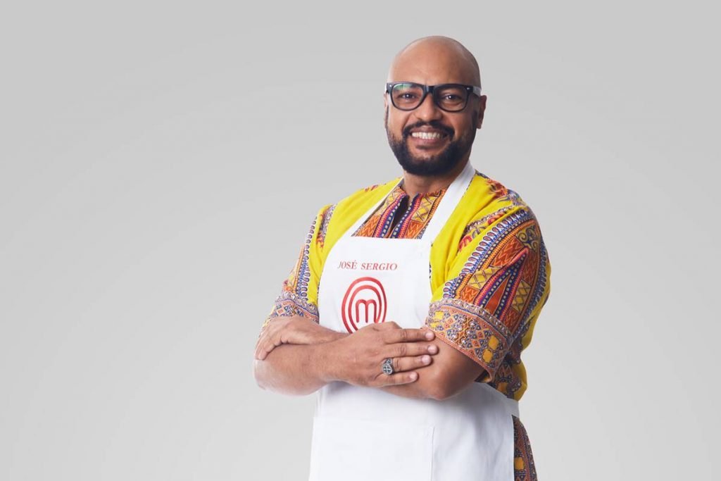 Homem de óculos de camisa de manga curta posa com avental do Masterchef de braços cruzados e sorri