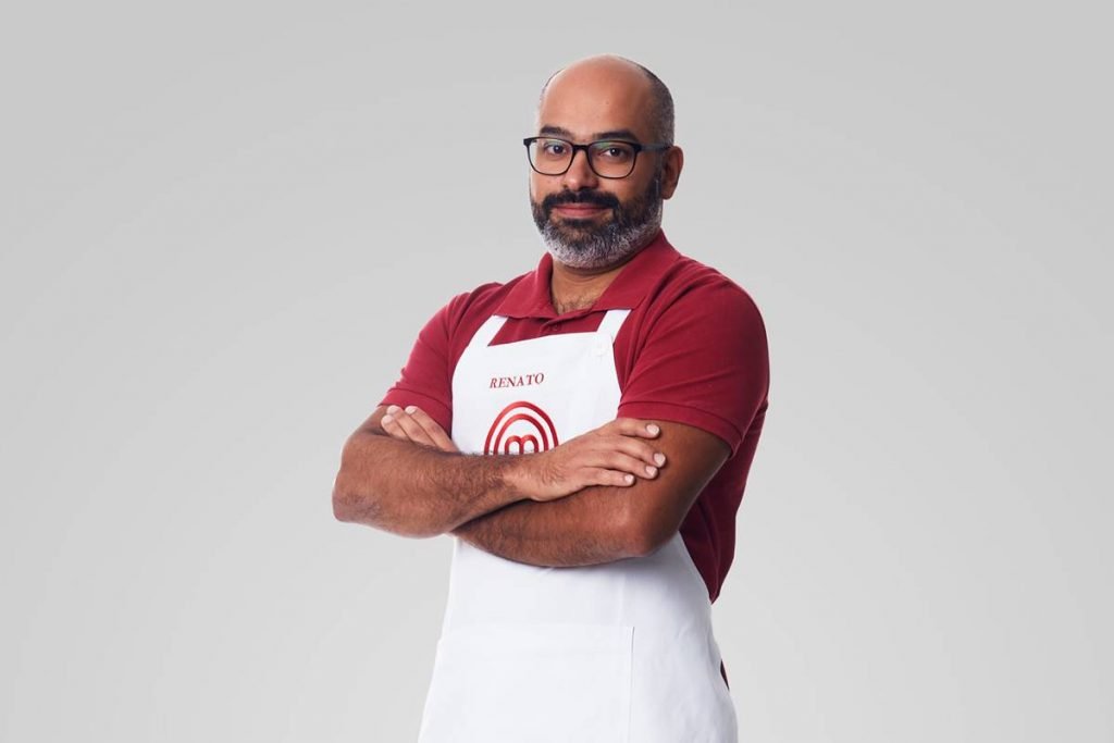 Homem de camiseta vermelha e óculos posa com avental do Masterchef de braços cruzados e sorri