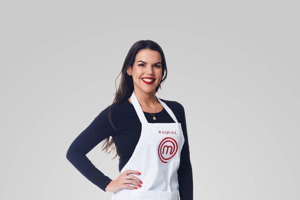 Mulher posa de blusa preta de manga longa com avental do masterchef e mão na cintura sorrindo