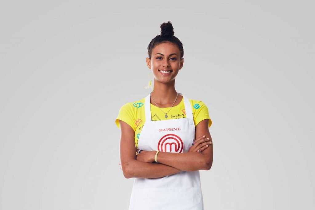 Mulher de blusa amarela posa com avental do Masterchef de braços cruzados e sorri