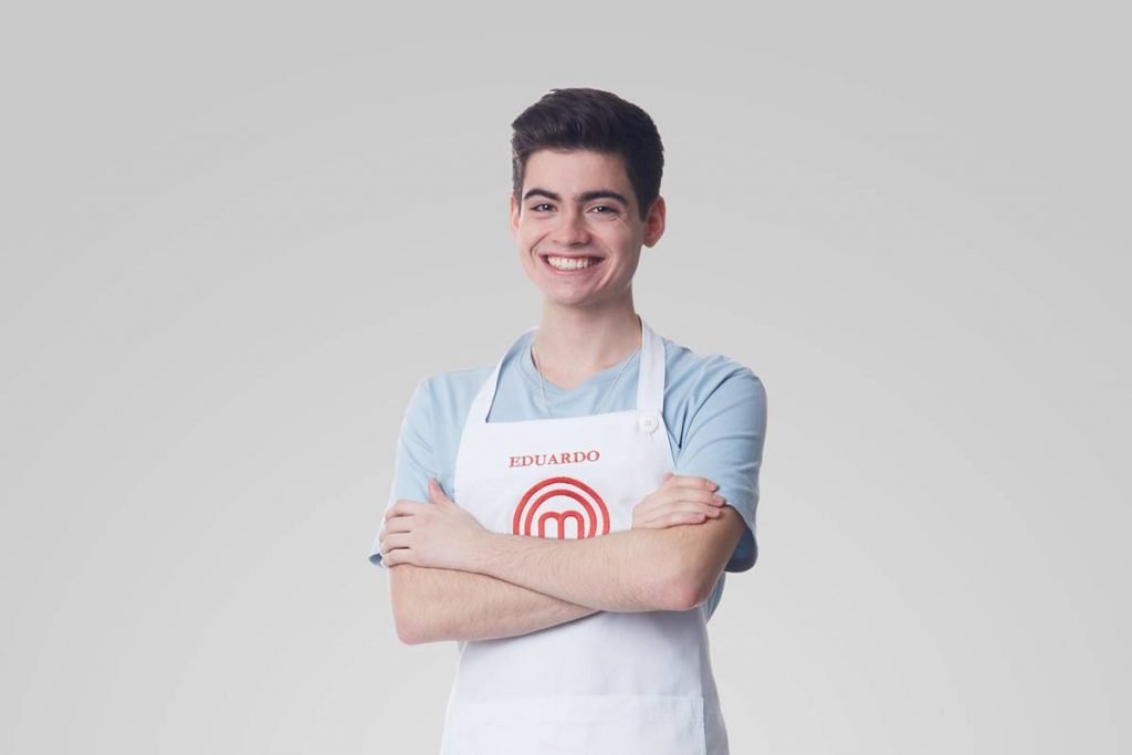 Homem de camiseta azul posa com avental do Masterchef de braços cruzados e sorri