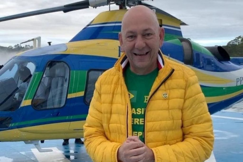 Fotografia colorida.  Luciano pose para foto com jaqueta amarela.  Ele está na frente de um helicóptero