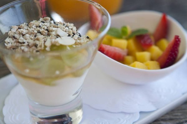 Copo com Iogurte caseiro com granola de quinoa e vasilha com frutas cortadas ao lado