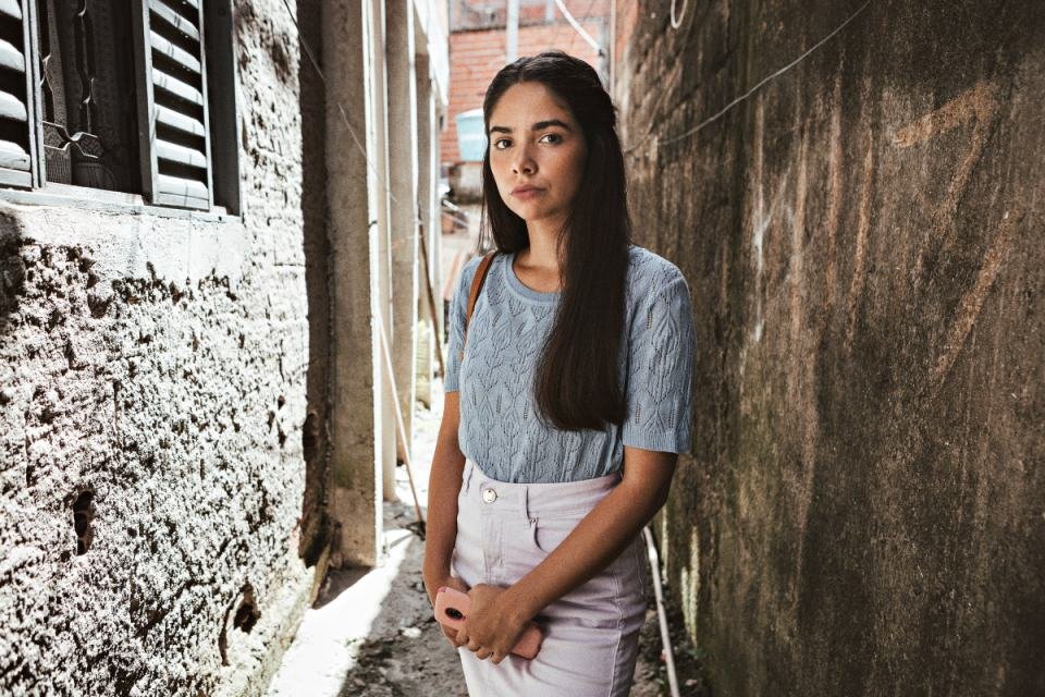 Bruna Mascarenhas, a Rita, de Sintonia. Ela está de blusa azul por dentro da calça branca e aparece em um beco na rua - Metrópoles