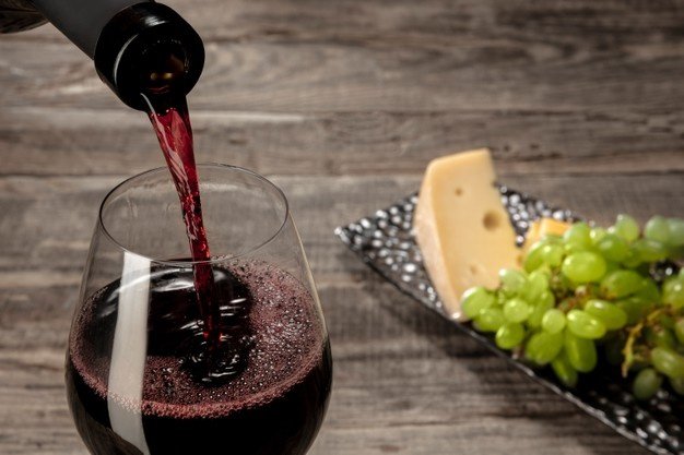 Na foto, uma garrafa de vinho está derramando o víquido roxo em uma taça, em cima de um fundo cinza, com uva verde e um pedaço dwe queijo
