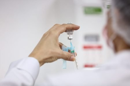 Uma mão com uma seringa tirando o líquido da vacina de um recipiente