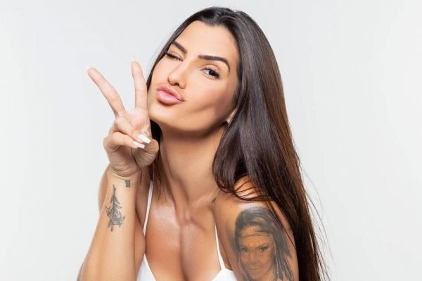Mari Ferrari aparece fazendo bico para a foto e levanta dois dedos para cima. Ela veste roupa branca