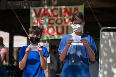 Adolescentes de camisa azul segurando cartão de vacina em frente a cartaz de posto de vacinação