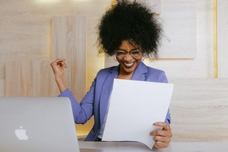 Mulher com papel na mão em frente a computador com sorriso de felicidade
