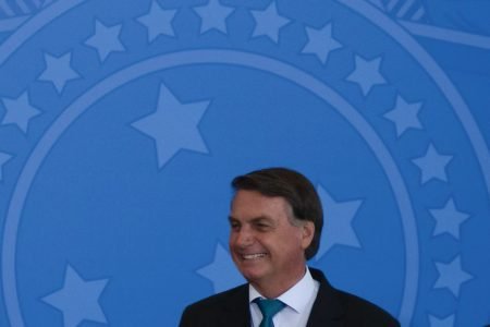 Presidente Jair Bolsonaro rindo; Ao fundo, escudo redondo azul-celeste, símbolo da república