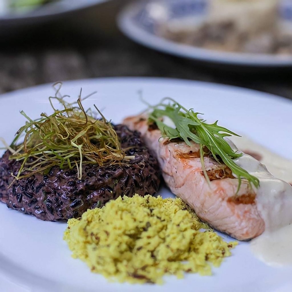 Salmão Quitinete do Universal, feito com salmão grelhado ao molho de queijo brie e coulis de morango, servido com risoto negro e farofa amarela