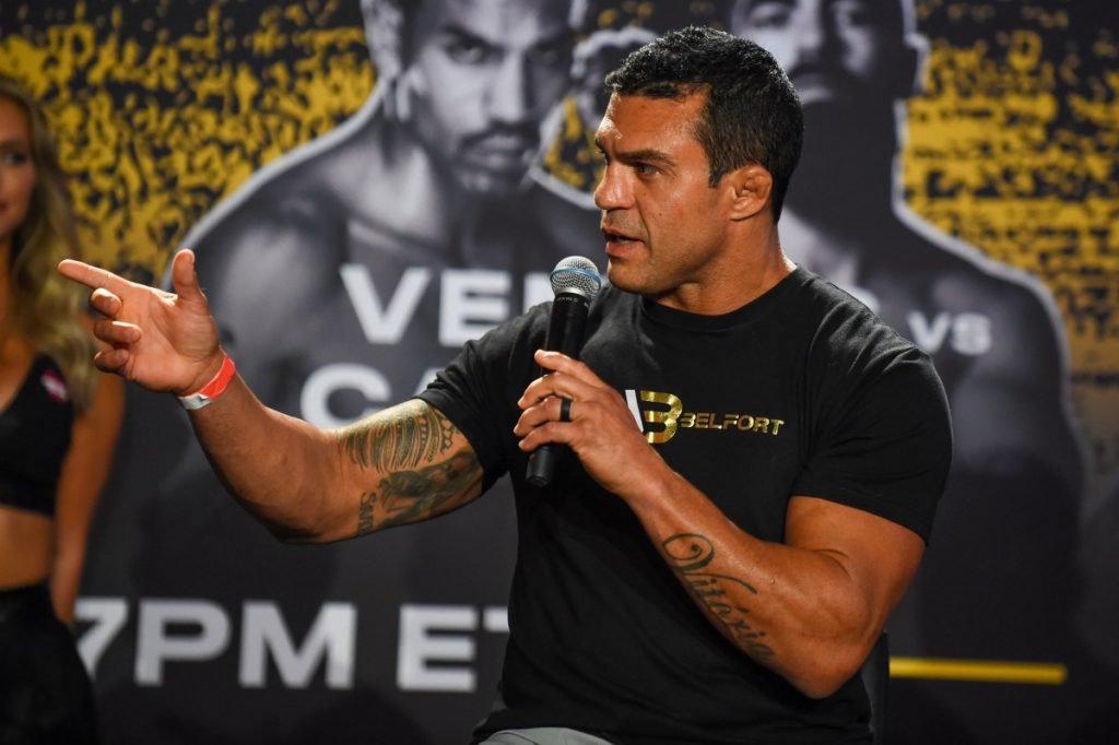 Vitor Belfort. O lutador está com um microfone na mão, camisa preta, tem tatuagens no braço e cara séria - Metrópoles