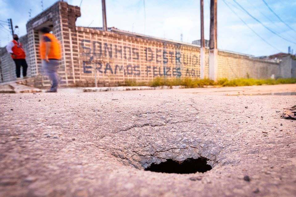 Imagem de enorme buraco em rua de Maceió. Ao fundo, um muro pichado com a seguinte frase: "Sonhos destruídos, éramos felizes"
