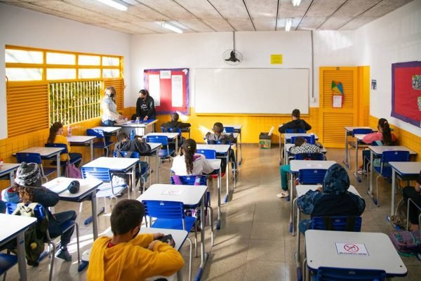 Educação. Imagem colorida mostra sala de aula com crianças sentadas em carteiras alternadas e professora na frente da sala - Metrópoles