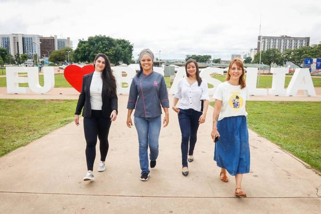 quatro mulheres caminhando com placa eu amo brasilia ao fundo