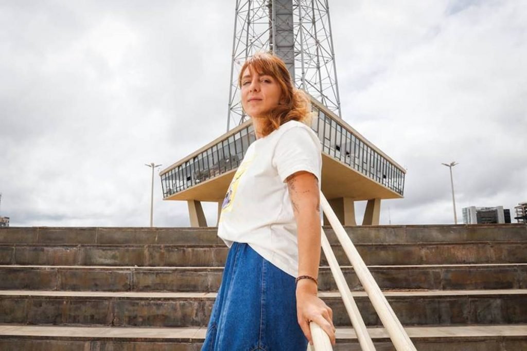 mulher de blusa branca apoiada de costas em corrimão na escada com torre de tv ao fundo