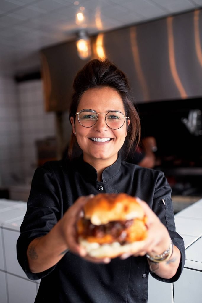 mulher de oculos e dolmã preta sorri e segura hamburguer na frente dela