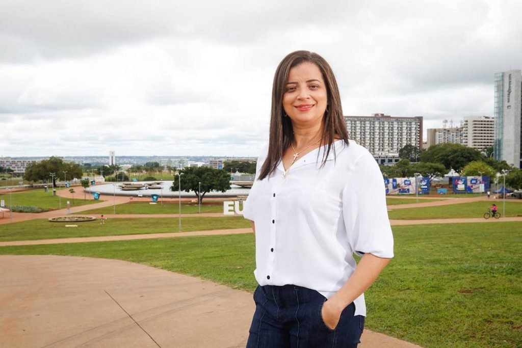 mulher de camisa branca e calça jeans com maos no bolso sorri para camera com fonte da torre de tv de fundo