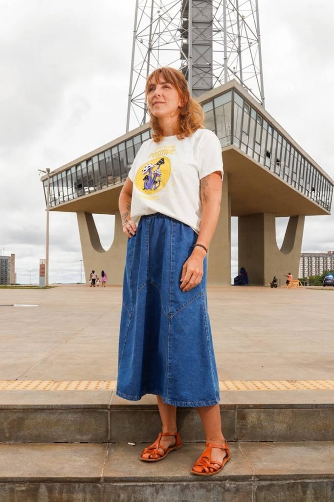 mulher de camiseta branca e saia jeans olha para o horizonte com torre de tv ao fundo