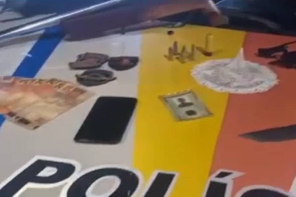 Em cima do capô de um carro tem um celular preto, uma identidade, e cédulas de dinheiro