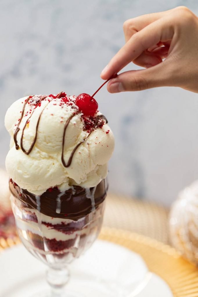 Fotografia de uma taça com camadas de bolo red velvet e creme de ninho com cream cheese, com uma bola de sorvete e uma cereja no topo. Do lado direito da imagem, uma mão segura a ponta do cabo da cereja
