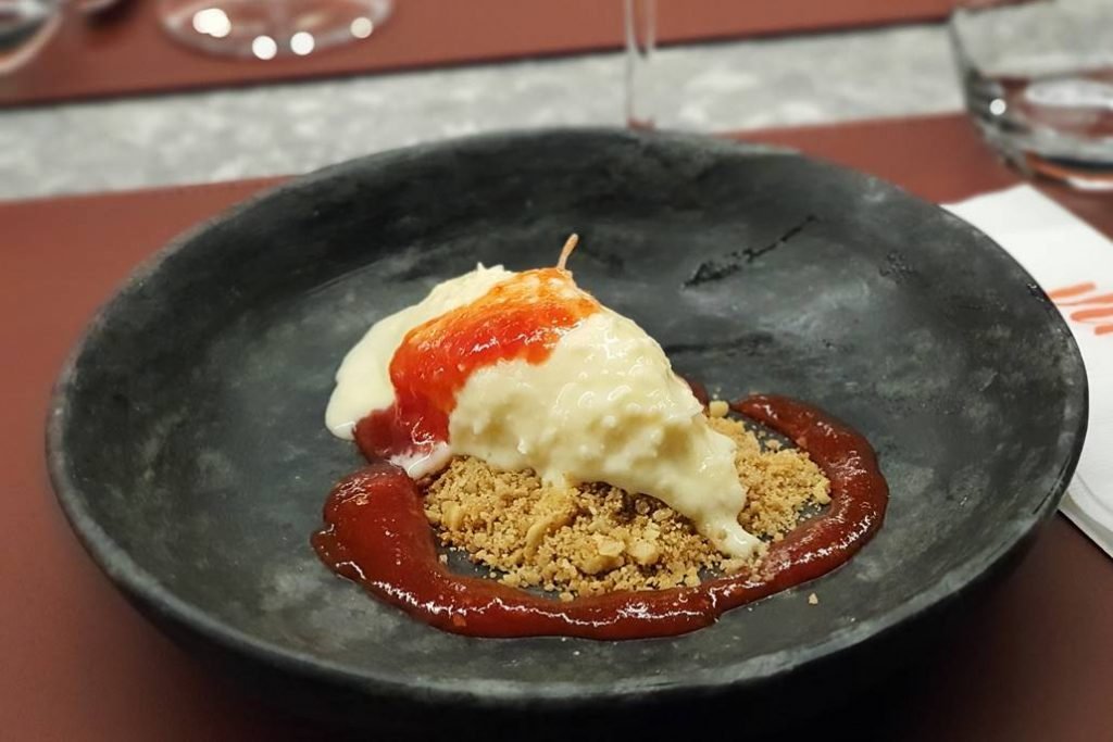 Fotografia de um prato cor chumbo com um pouco de sorvete cremoso sobre um crumble e geleia vermelha em volta do crumble e sobre o sorvete