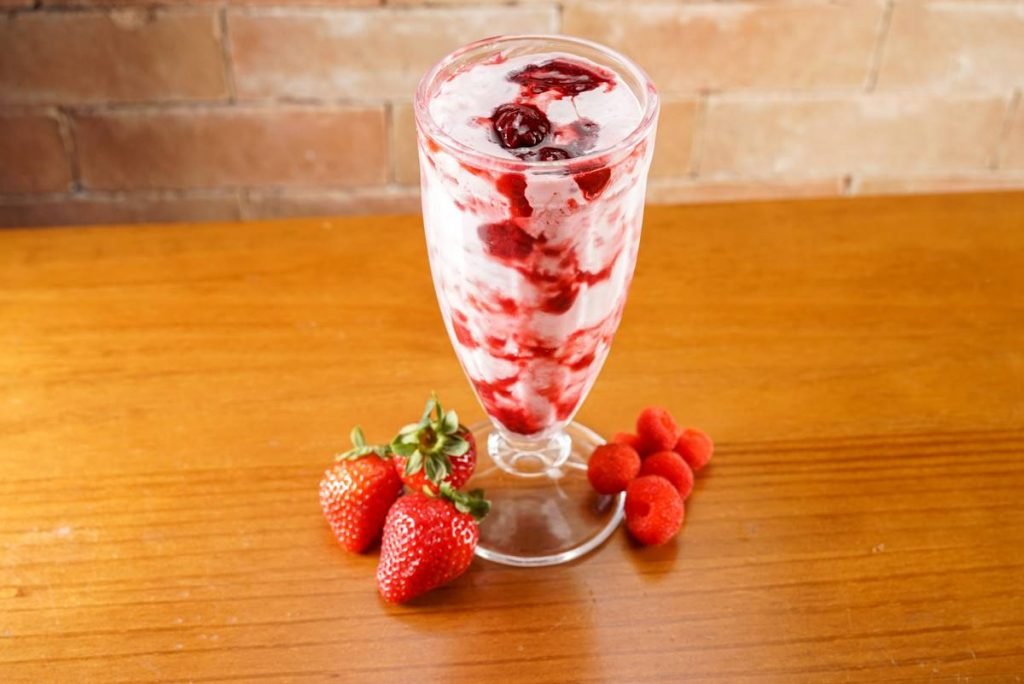 Fotografia de uma taça de milkshake com frutas vermelhas sobre uma mesa de madeira. Na base da taça, três morangos do lago esquerdo e cinco framboesas do lado direito