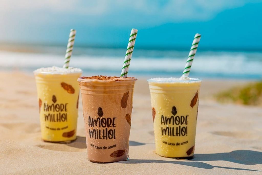 com uma montagem de areia e mar ao fundo, três copos de milkshake de milho estão dispostos lado a lado, com o meio mais para a frente. O do meio é marrom