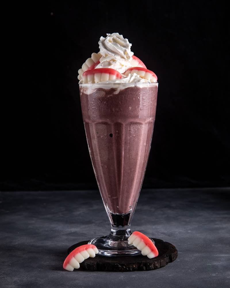 Na fotografia com fundo escuro, uma taça com milkshake de açaí, com chantilly no topo e balas de gelatina de dentadura