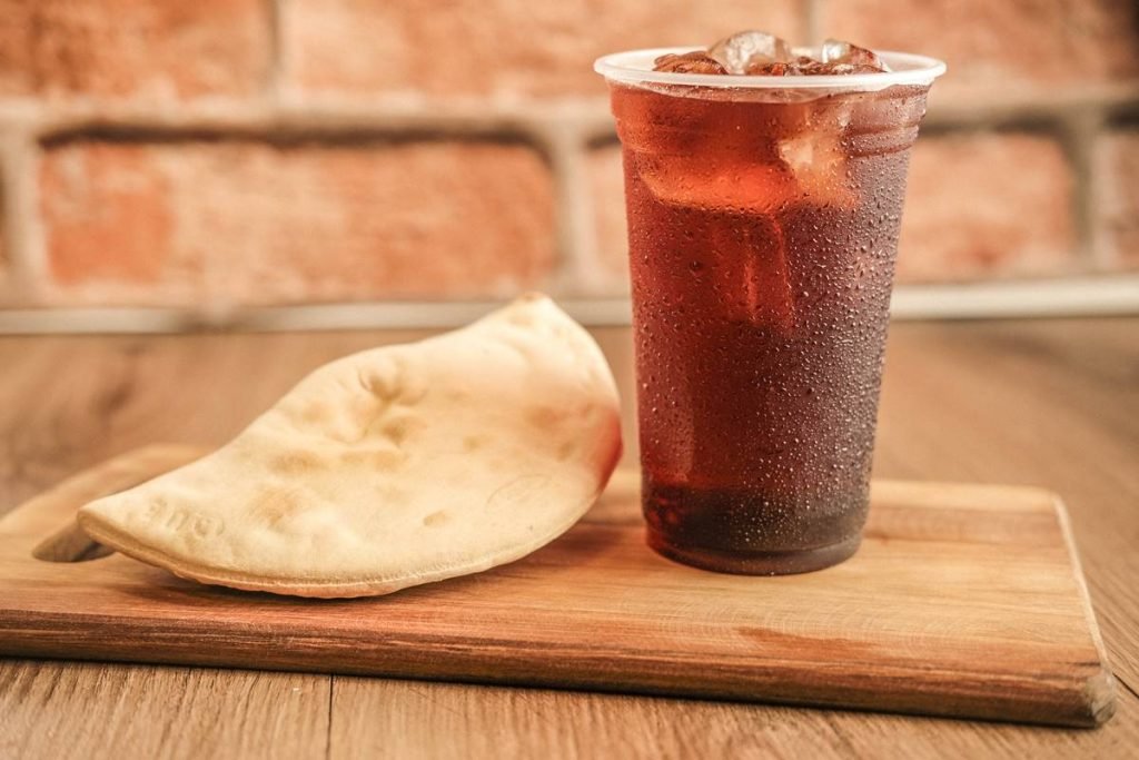 Na fotografia vemos uma tabua de madeira clara sobre uma mesa de madeira com um calzone e um copo plastico com chá e gelo