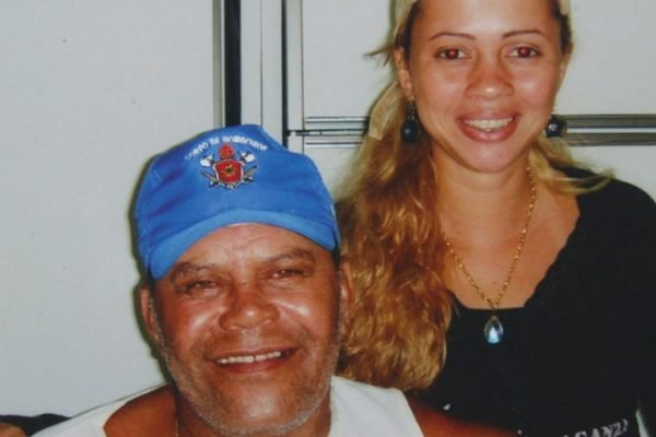 Fotografia colorida de Adriana Almeida, condenada pela morte de Rennê Senna. Ele, à esquerda da foto, usa boné azul. É um homem branco, velho, de barba rala e branca. Ela, à direita, é uma mulher branca, jovem, de cabelos loiros e longos. Os dois aparecem sorrindo, se abraçando.
