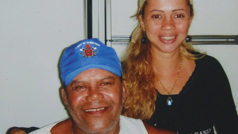 Fotografia colorida de Adriana Almeida, condenada pela morte de Rennê Senna. Ele, à esquerda da foto, usa boné azul. É um homem branco, velho, de barba rala e branca. Ela, à direita, é uma mulher branca, jovem, de cabelos loiros e longos. Os dois aparecem sorrindo, se abraçando.