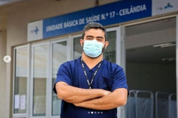 Homem de blusa azul e braços cruzados em frente a um centro de saúde