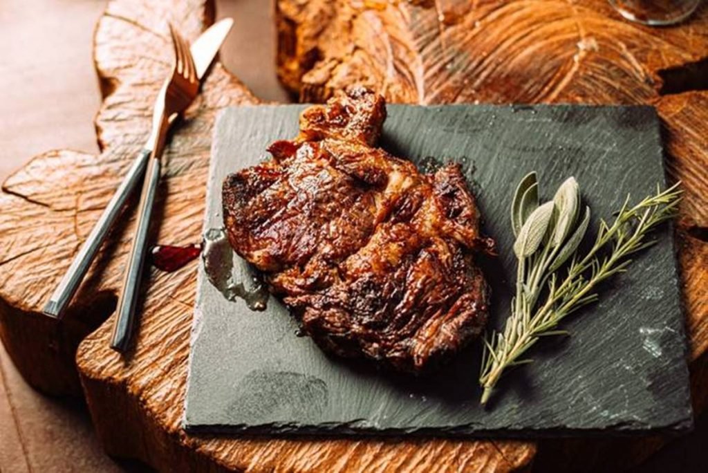 Na fotografia, um tabua cinza sobre uma mesa de madeira. Na tábua, um pedaço de bife cnacho assado com ervas do lado e um garfo e uma faca do lado da tabua, à esquerda