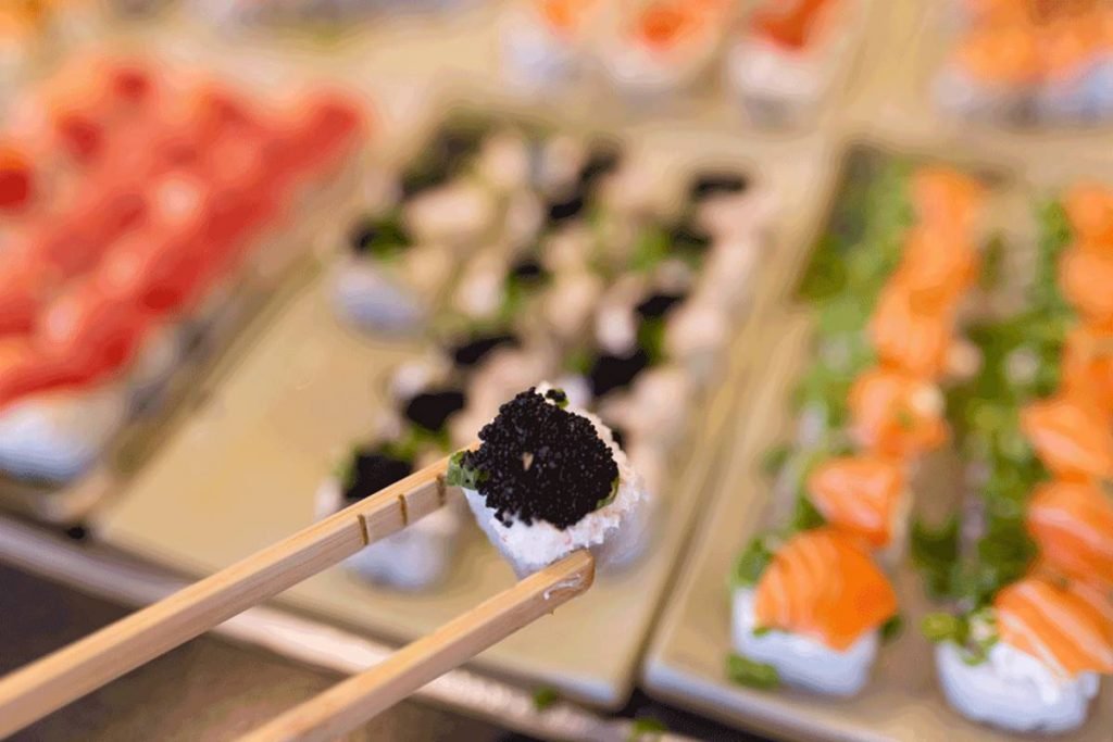 Na foto existem dois planos. No primeiro plano é possível ver um hashi segurando um sushi com patê de kane e ovas de peixe. Ao fundo, aparecem bandejas de sushis com salmão e ovas, mas embaçados.