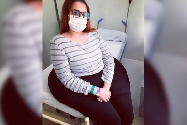 Imagem colorida com a mulher Karina Rodine sentada em uma cama. Karine vive em Curitiba, no Paraná, e tem um tumor de 40Kg