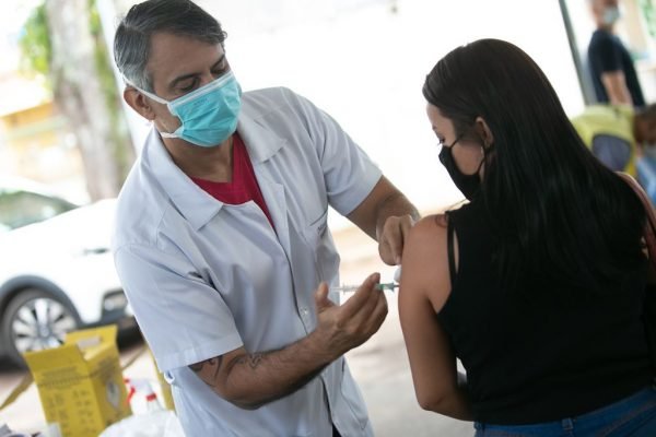 Enfermeiro aplicando vacina em menina de cabelo preto e blusa escura