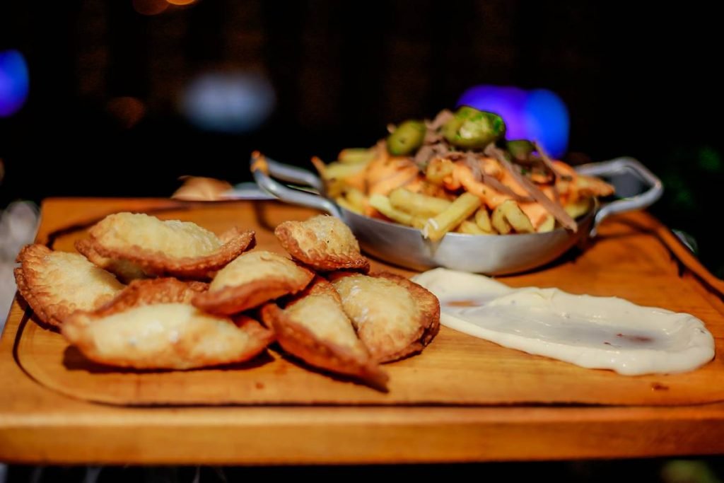 fotografia de tabua de madeira com uma porção de pasteis fritos a esquerda e um pouco de molho branco a direita. No fundo da bandeja, uma panelinha com batatas fritas cobertas de cheddar e azeitonas