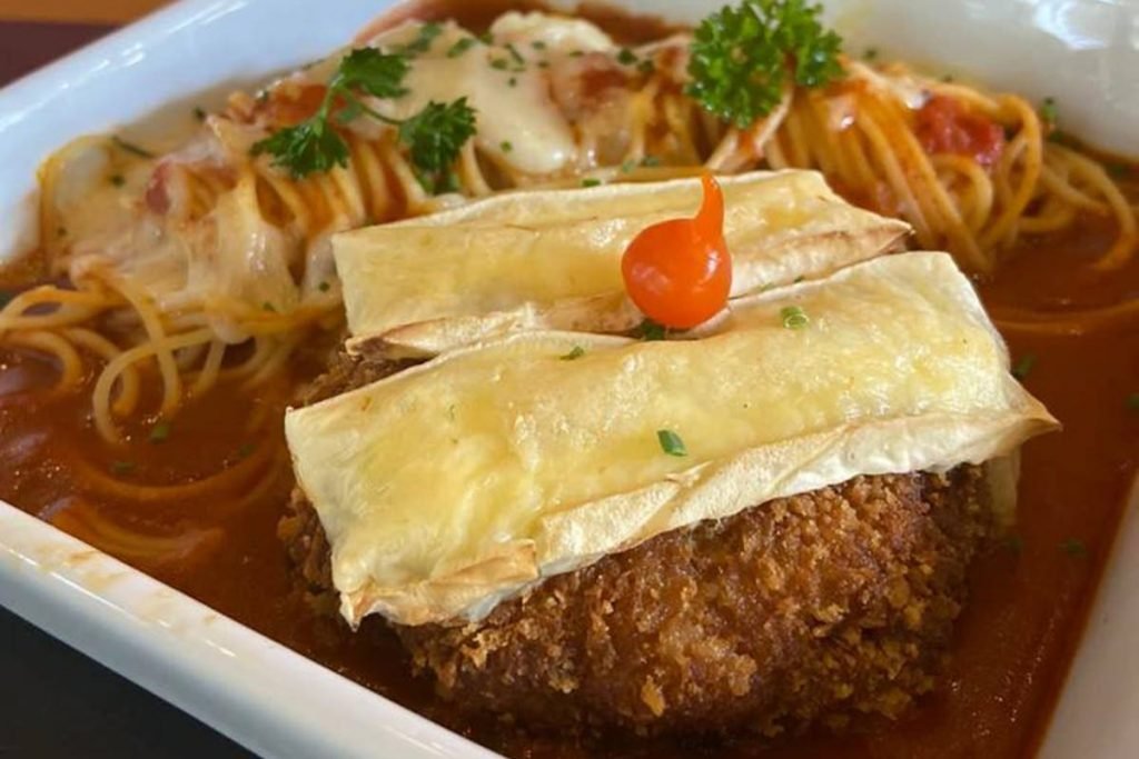 Fotografia de um prato quadrado com polpetone coberto com duas fatias de queijo brie e uma pimenta biquinho numa cama de molho de tomate, com espaguete enrolado ao lado e com folhas de salsa por cima