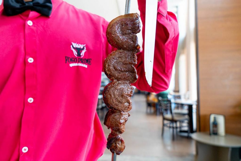 Na foto aparece o peito de um homem de camisa rosa com a logo da Fogo Prime. Ele segura um espeto de carne assada e uma faca