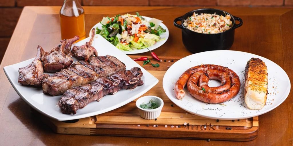 na foto, uma tabua de madeira com um prato de carnes a esquerda, um prato com linguiça e pao de alho a direita, um prato de salada ao fundo esquerdo e uma panelinha de arroz no fundo direito
