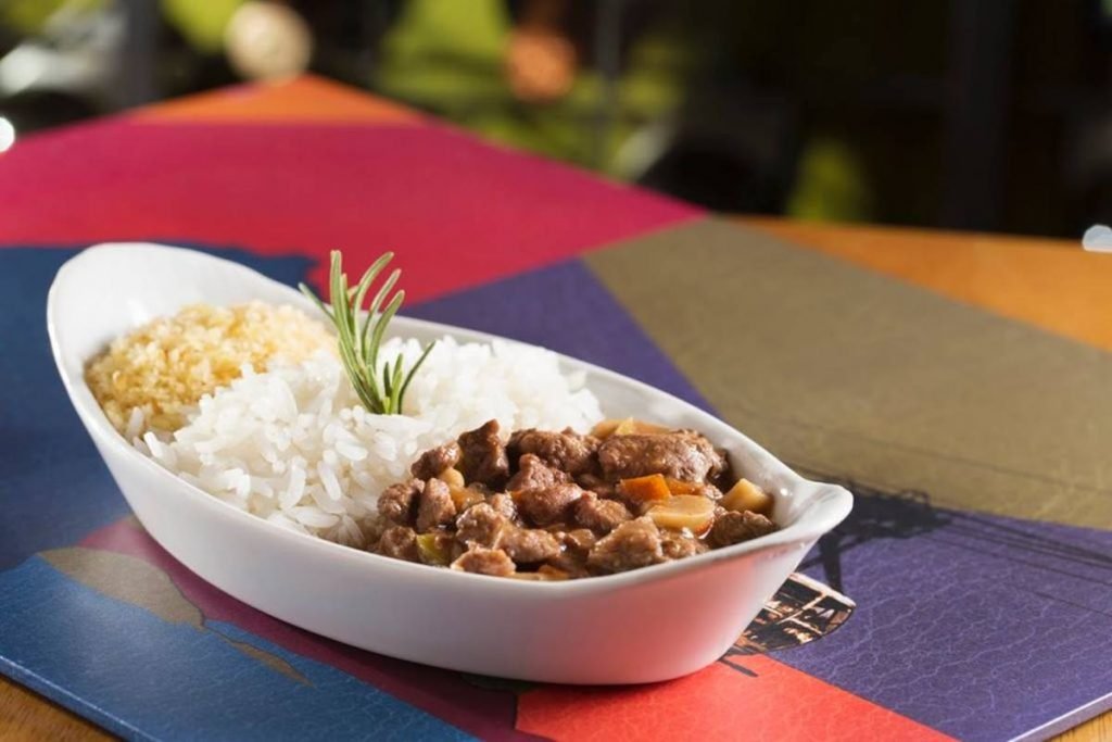 Na foto, uma mesa colorida com formas geométricas e uma vasilha de ceramica brnaca com picasinho, arroz branco e batata palha
