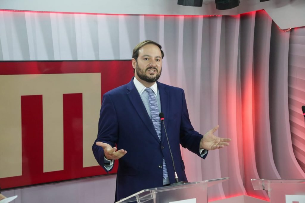 Homem de terno azul, camisa branca e gravata azul em frente a um púlpito falando em um microfone com as mãos para cima. Atrás um cenário branco e vermelho com o logotipo do Metrópoles, a letra M grande