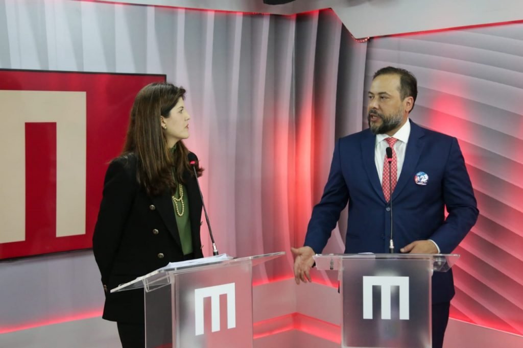 Mulher e homem um de frente para o outro. A mulher vestida de preto e verde e o homem vestido de terno azul