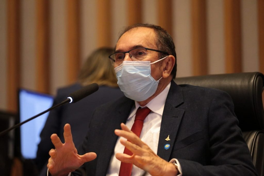 Homem de terno, gravata vermelha e máscara falando no microfone