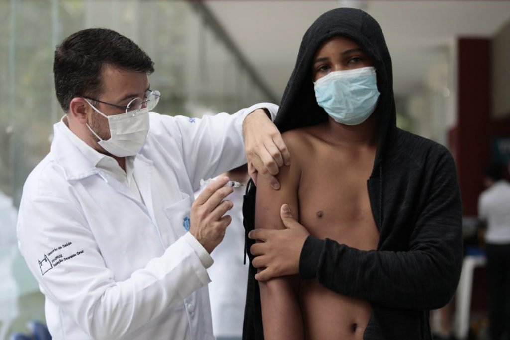 Secretário de Saúde do Rio, Daniel Soranz, aplicação vacina em um adolescente.  O médico é um homem branco, de jaleco branco, com óculos e máscara branca.  O jovem é negro, com um casaco preto e de máscara azul