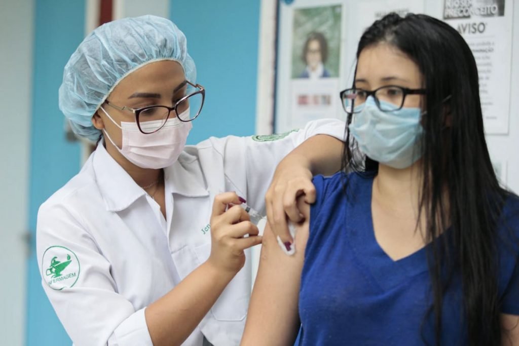 Cariocas com vacina da Covid-19 em atraso recebem imunizante. Na foto, uma jovem branca, de camisa azul, cabelos longos preto, óculos de grau preto e máscara azul